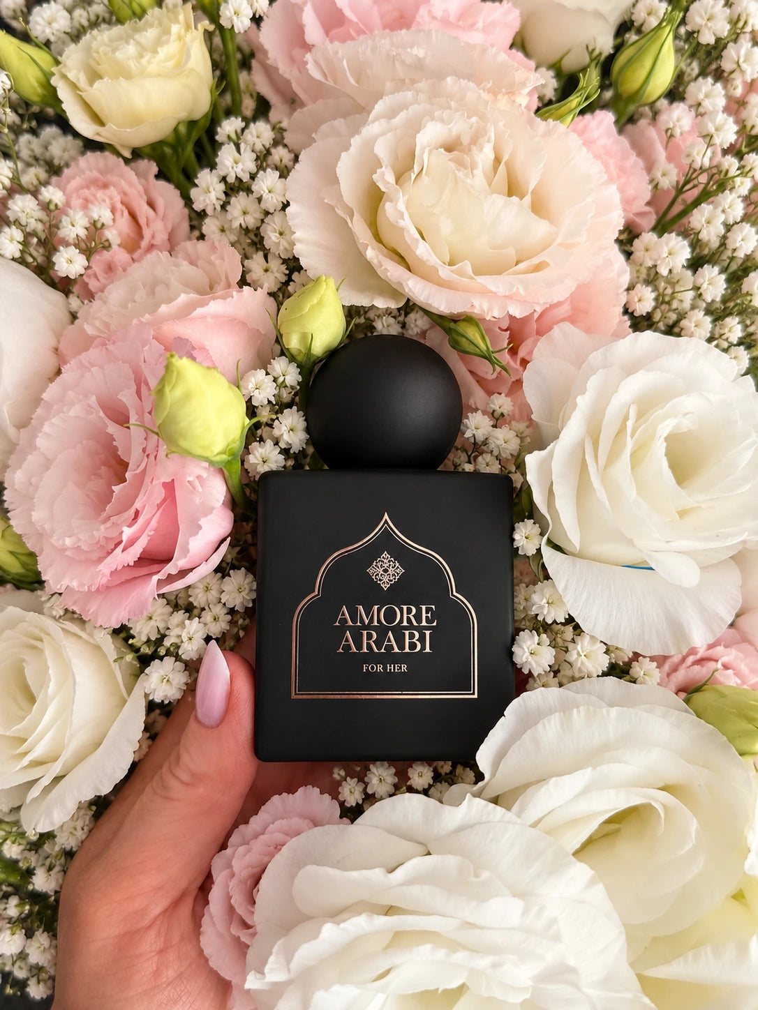 Czarny flakon Amore Arabi For Her trzymany w dłoni pośród białych i pudroworóżowych kwiatów